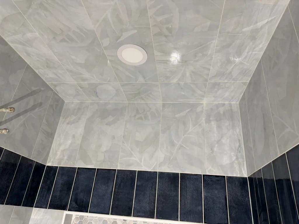 Looking up at a fully tiled shower ceiling in Manitou Springs, CO, featuring large-format light grey botanical pattern tiles, bordered by dark navy vertical accent tiles by XCEL TILE.