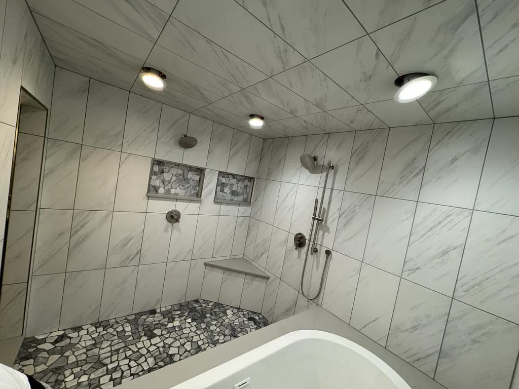 High-end wet room in the Broadmoor, CO, featuring white marble-look porcelain wall tiles, recessed niches, a corner bench, and a natural stone pebble floor by XCEL TILE.