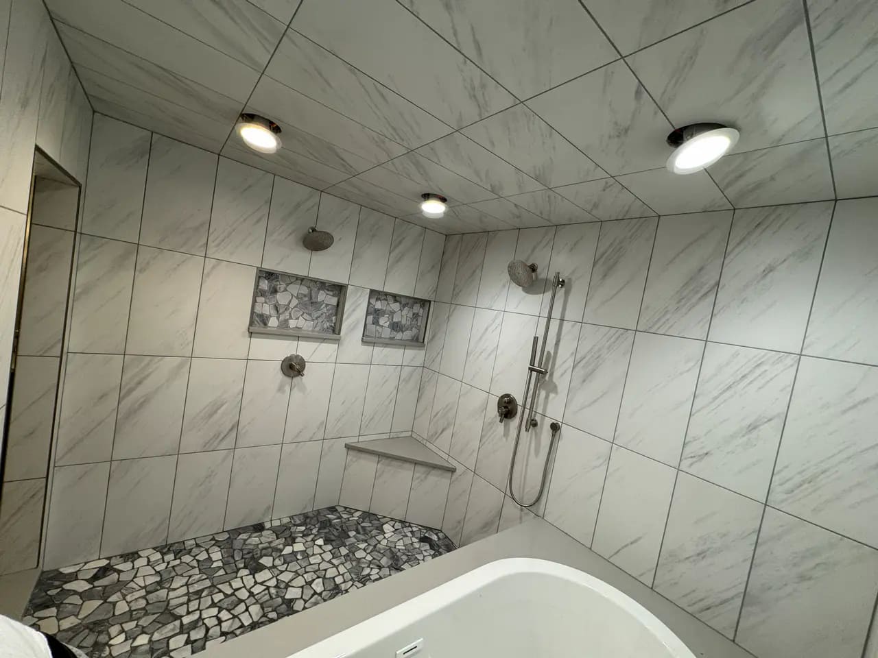 High-end wet room in the Broadmoor, CO, featuring white marble-look porcelain wall tiles, recessed niches, a corner bench, and a natural stone pebble floor by XCEL TILE.