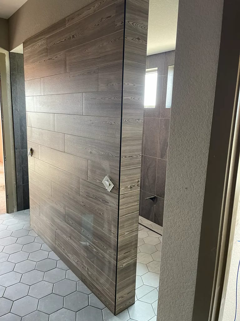 Custom bathroom partition wall in Castle Rock, CO, completely wrapped in horizontally stacked wood-look tile, contrasting with a light grey hexagon floor tile by XCEL TILE.